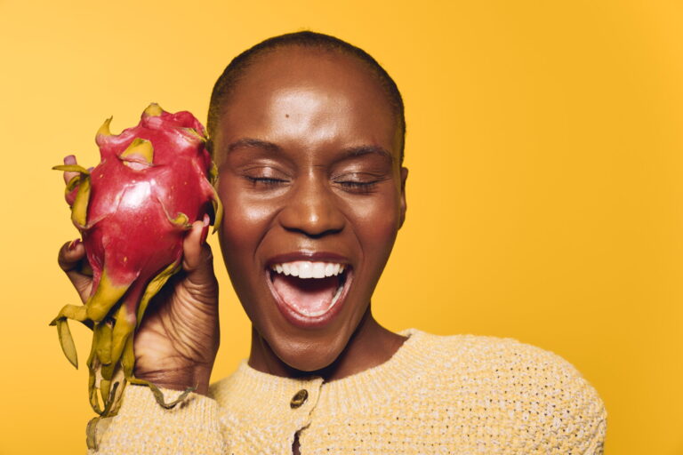 Smiling dark skinned African woman holds fresh dragon fruit near face with eyes closed. Yellow background and beige sweater create vibrant and joyful studio portrait. Concept of health and happiness.